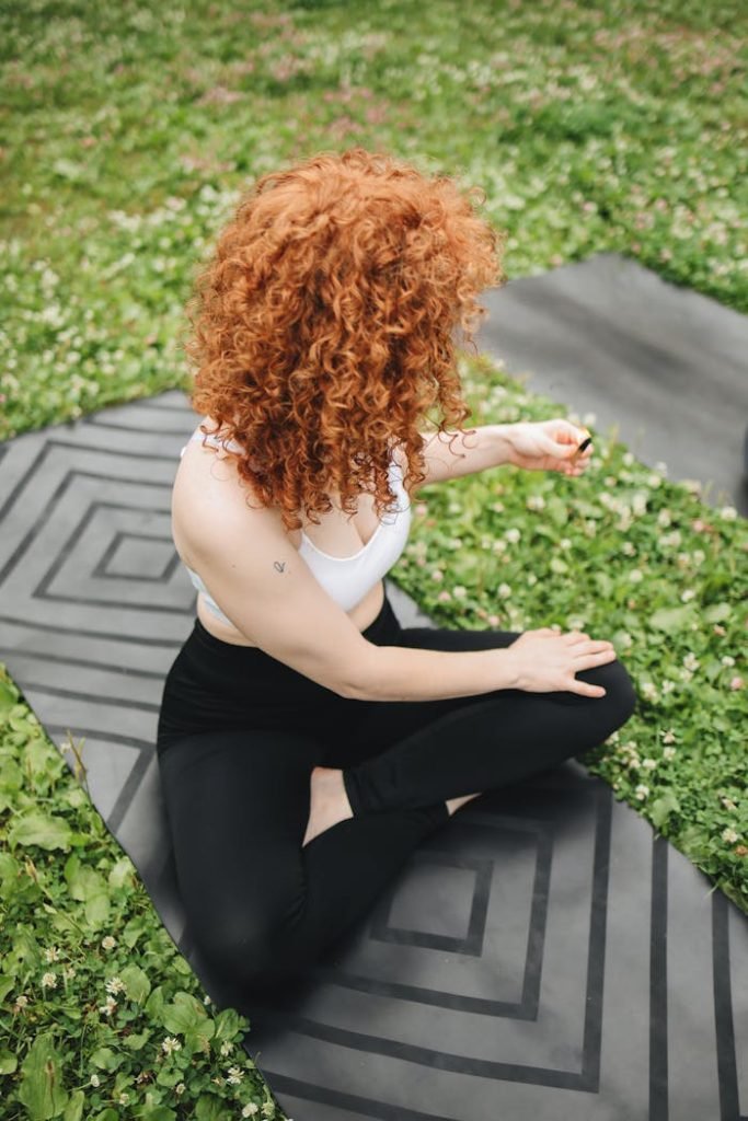 A woman with curly red hair sits on a yoga mat outdoors, meditating in a serene environment.